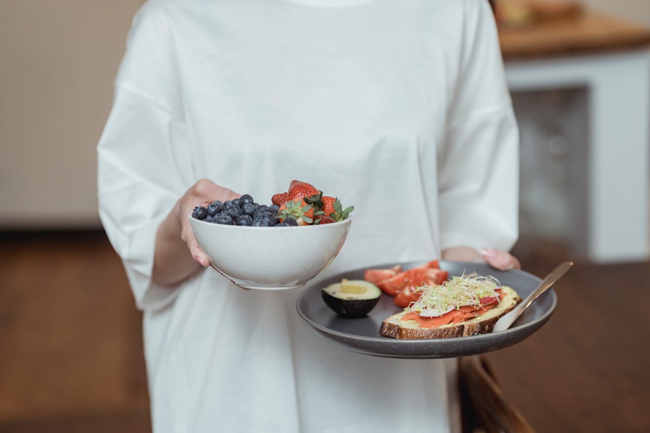 Person holding a bowl of fresh berries and a plate with avocado toast, displaying a healthy breakfast spread.